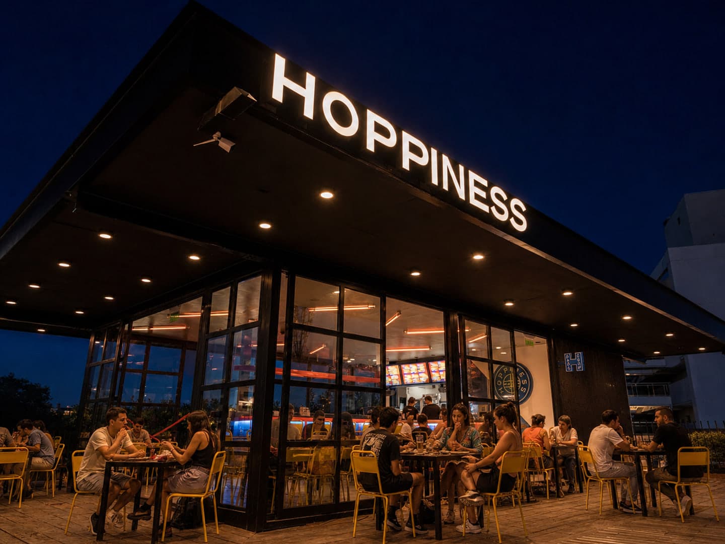 Exterior nocturno de Hoppiness Club Manantiales, con el salón vidriado y la terraza llena durante el servicio.