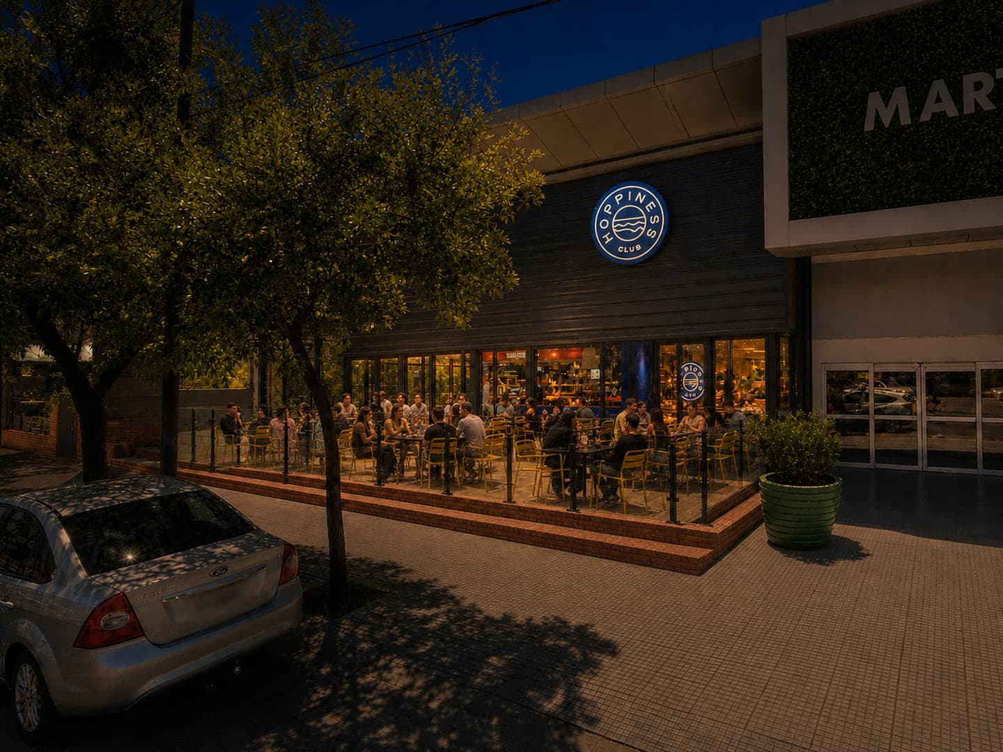 Visualización nocturna del futuro Hoppiness Club en Martinolli Mall, con la terraza al aire libre en funcionamiento y el cerramiento perimetral de vidrio.
