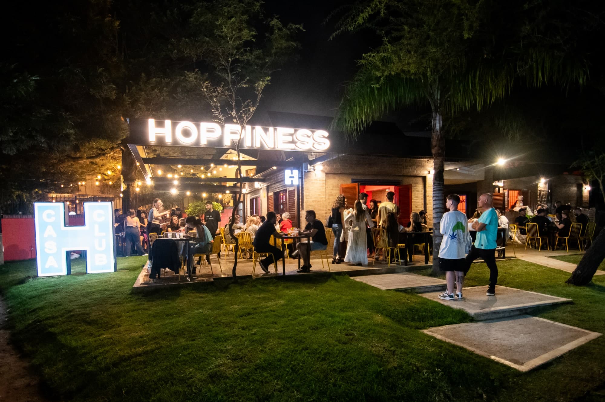 Exterior nocturno de Hoppiness Club Villa Allende, con el jardín y la terraza llenos de gente.